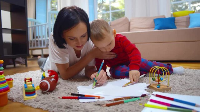 Caring young mum or nanny helping cute kid drawing picture with pencils lying on floor. Realtime