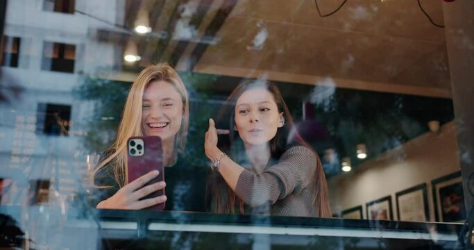 Young women taking selfie in restaurant. Low angle handheld shot of young brunette showing tongue while taking selfie with happy girlfriend behind glass in restaurant