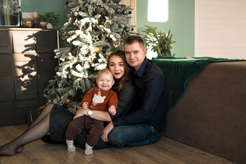 Family with baby are sitting on  floor by Christmas tree and smiling sweetly.