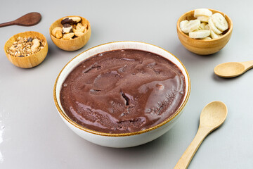 Brazilian açai in a white bowl, additional banana, granola and nuts in small bamboo bowls. Cashew and Pará nuts. Top view. Gray background. Selective focus