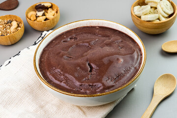 Brazilian açai in a white bowl, additional banana, granola and nuts in small bamboo bowls. Cashew and Pará nuts. Top view. Gray background. Selective focus