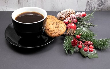 Coffee in a black cup with glazed cookies, red holly berry and spruce branch on a black background. Christmas card with copy space.