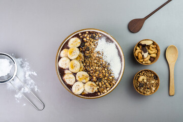 Brazilian açai in a white bowl with banana, powdered milk, honey, granola and nuts in small bamboo bowls. Cashew nuts and Pará. Top view. Grey background. Selective focus