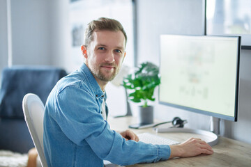 Young handsome IT support man in denim shirt . Attractive bearded caucasian male freelance software engineer sitting at working place in office hardly working using computer. High quality image