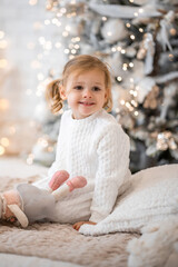 Fototapeta premium Very nice charming little girl blonde in white dress sitting on a child's bed and laughs on the background of Christmas trees in bright interior of the house
