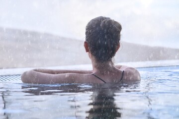 Woman enjoying view, relaxing in outdoor thermal pool on a winter day while it is snowing. Snowy mountains on the background. - obrazy, fototapety, plakaty