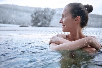 Happy woman smiling, enjoying view, relaxing in outdoor pool on a winter day while it is snowing. Snowy mountains on the background. - obrazy, fototapety, plakaty