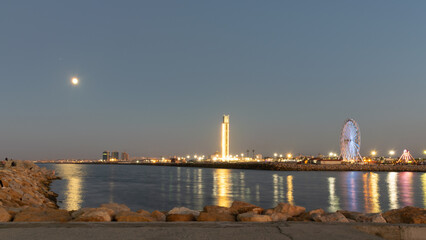 Obraz premium The great mosque and ferris wheel under moonlight seen from Sablette promenade jetty. Skyline view of Algiers city by night, water reflexion lights illuminated by colorful world biggest minaret.