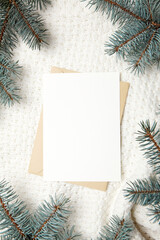 Fototapeta na wymiar Christmas greeting card mockup with envelope and fir tree branches on white winter knitted pullover background, top view