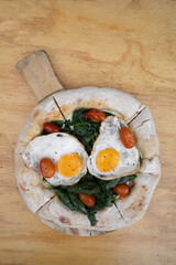 Top view of a pizza with mozzarella cheese, spinach, cherry tomatoes and fried eggs, on the wooden table.	