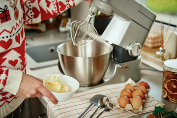 Woman in festive christmas sweater preparing dough for cookies at home kitchen. Female hands use electric mixer. Modern kitchen household appliances