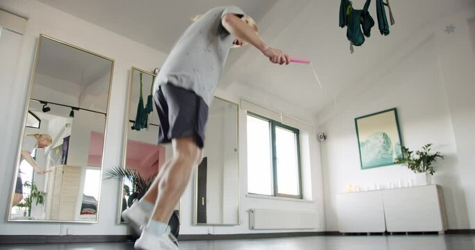 Sportsman doing tricks with jumping rope. Handheld tracking shot of energetic blond male athlete in sportswear doing tricks with jumping rope during fitness workout in sunlit gym