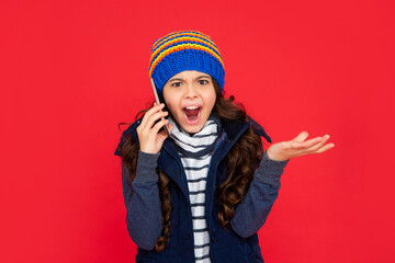 unpleased kid talking on phone in hat on red background. teen girl in down vest with smartphone.