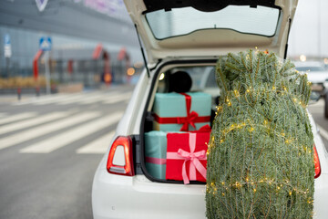 Wrapped Christmas tree decorated with garland near car trunk full of gift boxes outdoors. Concept...