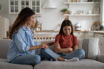 Positive young loving woman mother talking to angry offended daughter teenage girl, smiling mom trying to cheer up teen kid while sitting together on sofa, adolescent child wont speak with parent