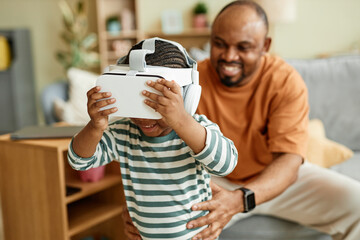 Portrait of cute black kid wearing VR headset and enjoying virtual reality entertainment for children with father in background