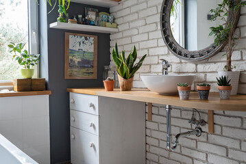 Modern bathroom in industrial style. Loft interior of bathroom with brick wall, countertop with ceramic washbasin and round mirror. Houseplant and spa at home.