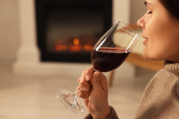 Young woman with glass of wine relaxing near fireplace at home, closeup. Space for text