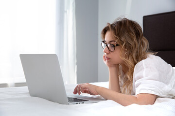  A beautiful girl with glasses is lying on a white blanket in the morning light, chatting, online, typing on a computer keyboard - the concept of a social network, online shopping.