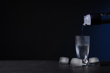 Pouring vodka from bottle into shot glass on black table against dark background. Space for text