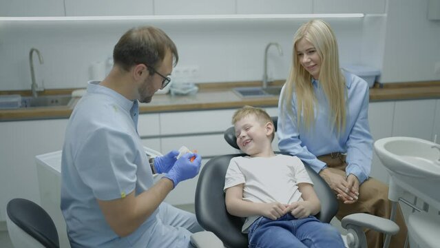 Male dentist shows mock-up of jaw to boy and his mother how to brush their teeth properly in playful way. Child sits in dental chair and claps his hands. Slow motion