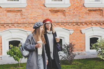 Fototapeta premium Young female best friends talking to each other discussing interesting themes during free time hipster girls joking and having fun together enjoying recreation with coffee to go on urban background