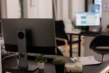 Multiple workstations with lighted monitors on desks, indoor plants and landline phone on the table. Empty office of financial company before starting the daily work day.