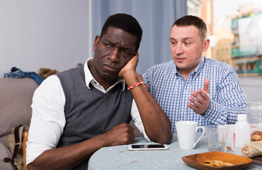 Two sad interracial men sitting at table after conflict at home interior