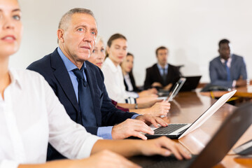 Fototapeta na wymiar Mature senior white male manager attending business meeting in conference room and intently listening to colleague's report together with coworkers sitting in row