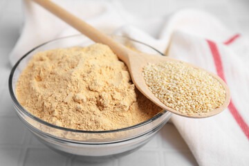 Sesame flour in glass bowl with wooden spoon of seeds on white table, closeup