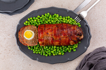 Top view of baked meatloaf made of ground meat, onion, carrot, stuffed with hard-boiled eggs, wrapped with bacon net, served with green peas. Easter food.