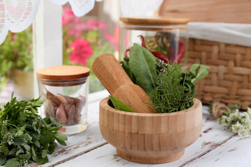 Mortar with pestle and fresh green herbs on white wooden table near window
