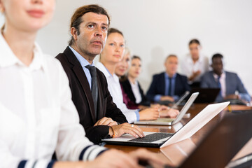 Focused mature white business man in formal suit sitting with colleagues in conference room and absorbedly listening 