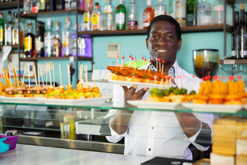 Smiling African American bartender standing with plates of pinchos in front of a pub counter. High quality photo