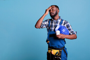 Exhausted construction employee drinking coffee during break. Overworked worker holding protective helmet wearing bibs while resting against blue background in studio shot.