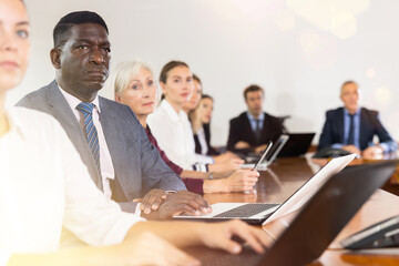 Fototapeta premium African-american businessman using laptop during conference in meeting room. Man sitting at desk with colleaugues.