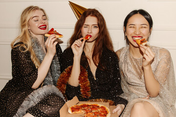 Image of three young multicultural women in sparkly dresses eating pizza sitting on floor....
