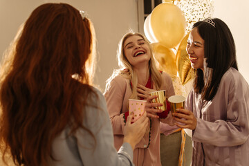 Happy young interracial girls in good company celebrating birthday in room with balloons. Pretty...