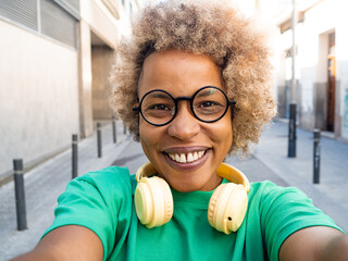 Perspective selfie of a happy african american woman with afro hair wearing glasses and headphones in the city 