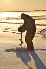 An ice fisherman drilling a hole at sunset