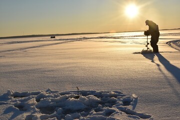 An ice fisherman drilling a hole at sunset  - obrazy, fototapety, plakaty
