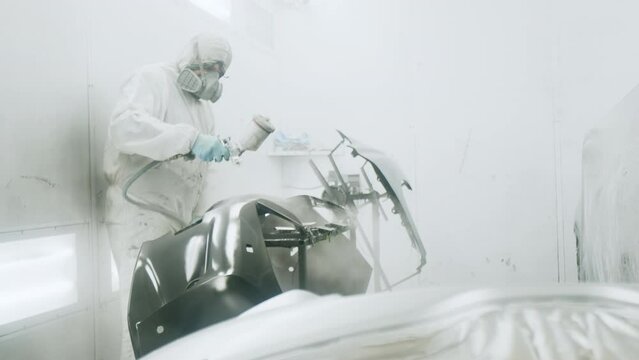 Detailed view of a worker in white, protective, suit. Professional, industrial painter wearing a mask while spraying car parts at the car service station. High quality 4k footage