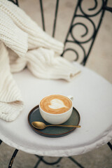 Cup of hot cappuccino on a chair near white knitted sweater.