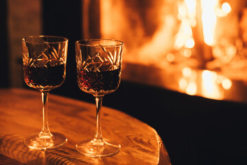 Two glasses of red wine on wooden table with cozy fireplace flame on the background.