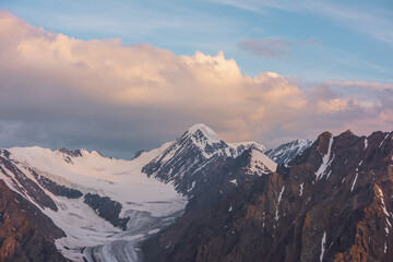 Obraz premium Dramatic aerial view to high snow mountain peak in early morning at dawn. Awesome scenery with sunlit snow mountains in cloudy sky at sunrise. Scenic landscape with large glacier in sunrise colors.