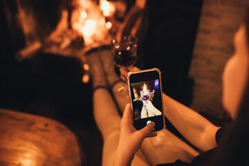 Young woman drinking red wine sitting by the fire in front of cozy fireplace, snapping herself using camera on smartphone.