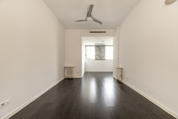 Empty living room of a house with a dark brown laminate floor, several cast iron radiators and a fan with gray blades on the ceiling