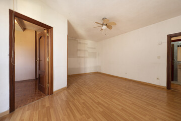 Empty living room of a house with wood-like laminate flooring, a fan with blades on the ceiling and dark wooden doors