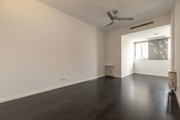 Empty living room of a house with a dark brown laminate floor, white cast iron radiators, a wall to wall window and a fan with gray blades on the ceiling