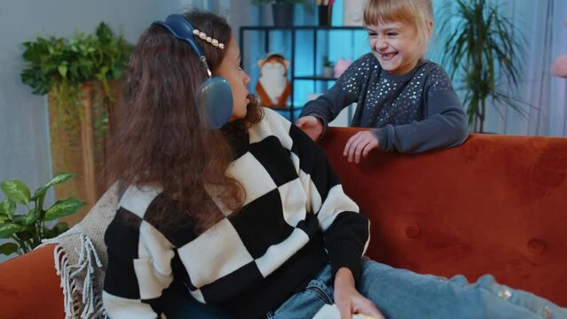 Little girl kid frightening her scared afraid sister child while relaxing and reading book. Game, prank, joke, Fools Day. Female siblings children or best friends at home playroom. Friendship family